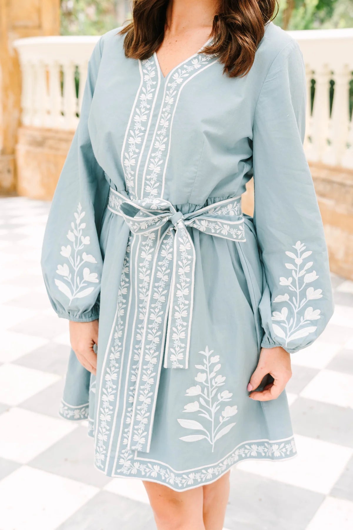 A woman wearing a dusty blue long-sleeve tunic dress with white floral embroidery and a matching tie belt, standing on a black and white checkered tile floor.