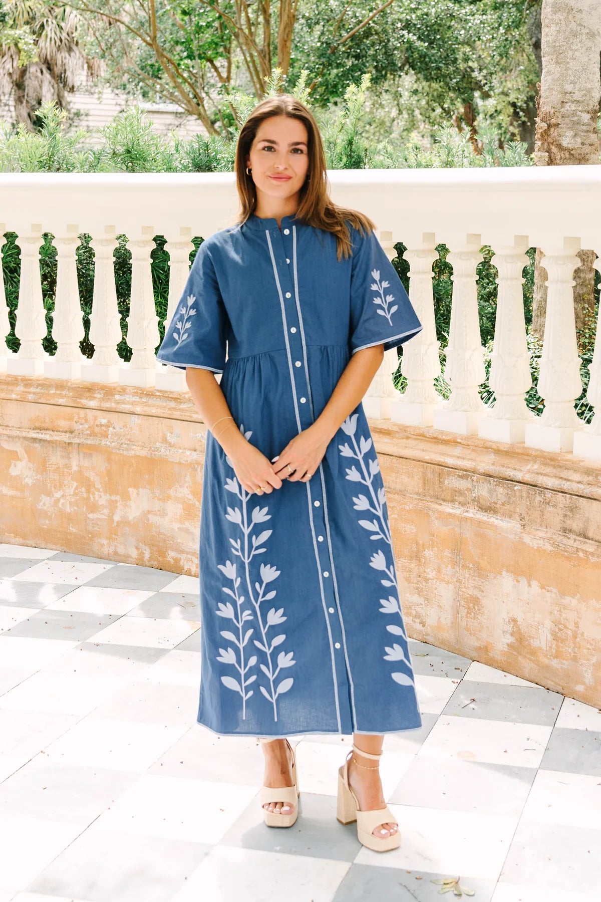 A woman wearing a blue midi dress with white floral embroidery, standing outdoors in front of a stone balustrade and tiled floor.
