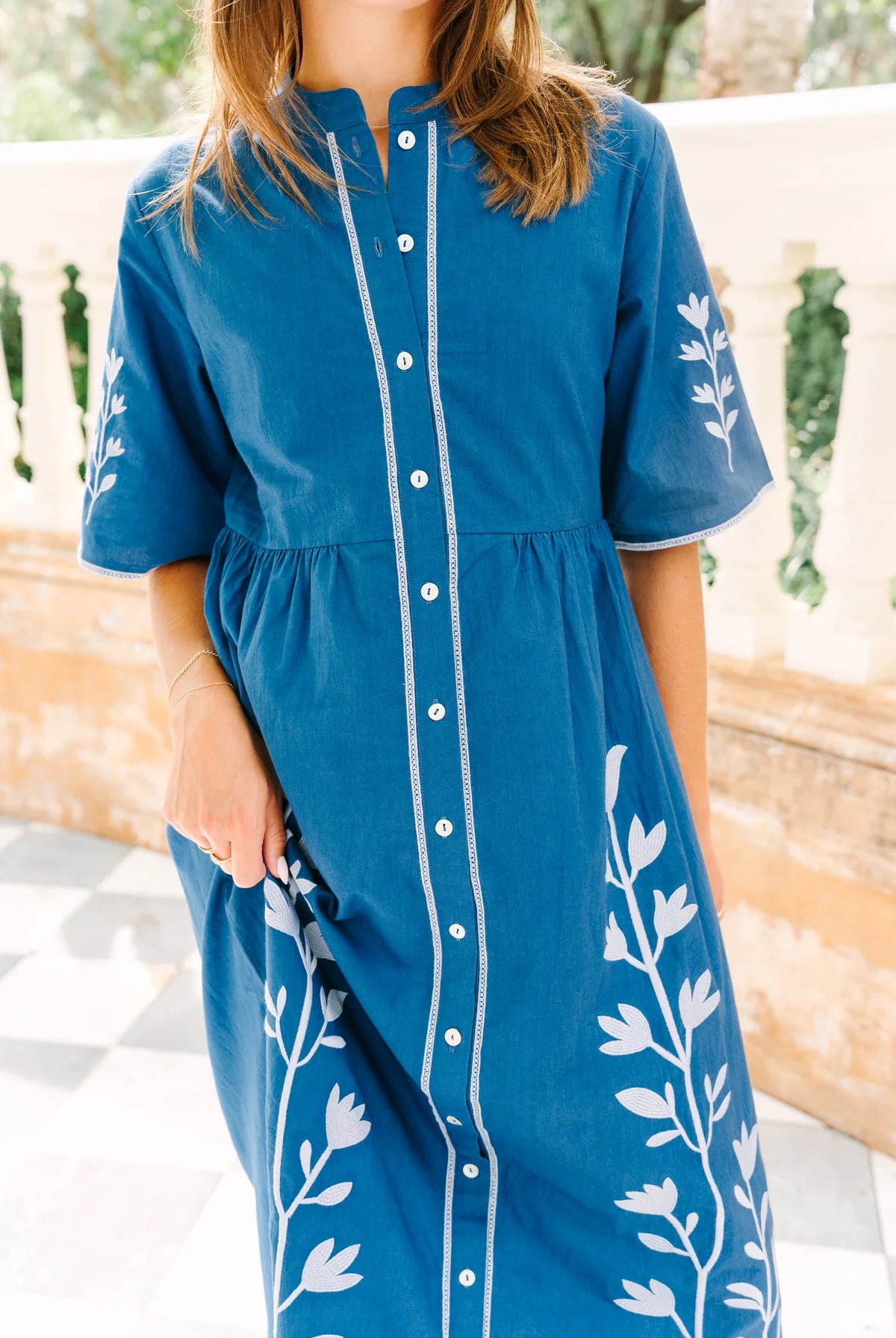 A woman wearing a blue short-sleeve button-front dress with white leaf embroidery on the sides and sleeves, photographed outdoors against a light stone wall.