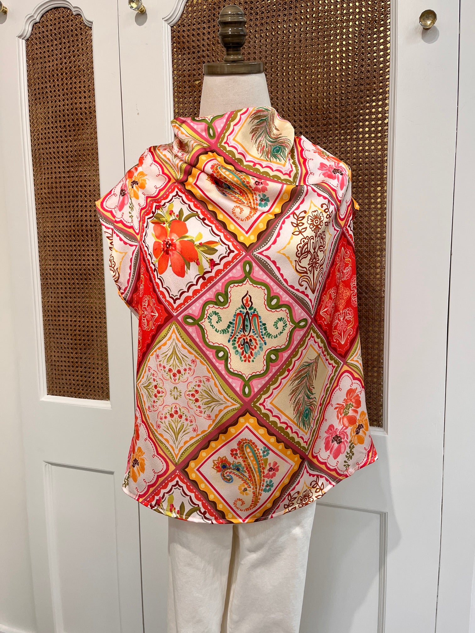 A folded textile item with a colorful patchwork-style floral and geometric print in red, pink, cream, green, and yellow, draped over a white chair in front of a white cabinet with woven inserts.