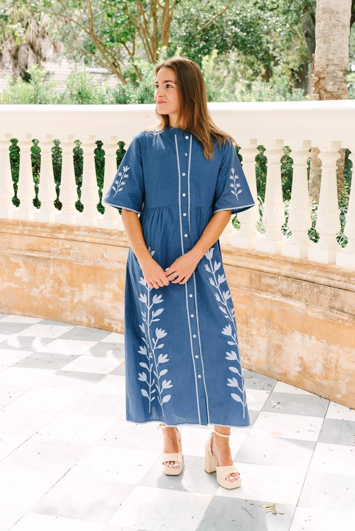 A woman wearing a blue midi dress with white floral embroidery and white buttons, standing on a tiled patio in front of a stone balustrade.