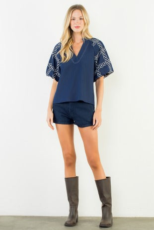 A woman modeling a navy blue short-sleeve top with white chain embroidery on the sleeves, paired with matching navy shorts and dark brown knee-high boots, against a plain white studio background.