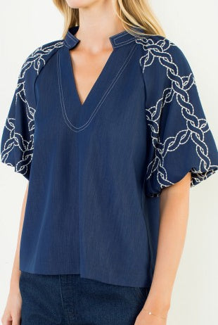 A woman wearing a navy blue blouse with a split V-neckline and short flutter sleeves featuring white embroidered chain motifs, paired with dark blue jeans against a plain light background.