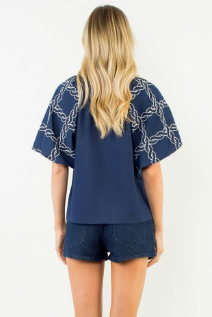 A woman viewed from the back wearing a navy blue short-sleeve top with white chain-link patterned sleeves and dark denim shorts, against a plain white background.