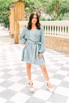 A woman modeling a light blue denim-style mini dress with white embroidery and a tie waist, standing on a gray and white checkered outdoor patio.