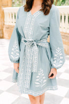 A woman wearing a dusty blue long-sleeve tunic dress with white floral embroidery and a matching tie belt, standing on a black and white checkered tile floor.