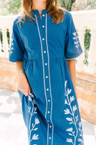 A woman wearing a blue short-sleeve button-front dress with white leaf embroidery on the sides and sleeves, photographed outdoors against a light stone wall.