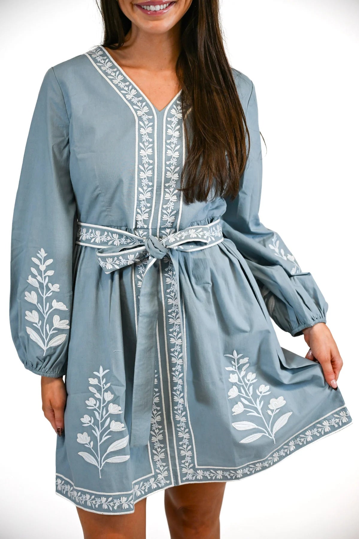 A woman wearing a dusty blue long-sleeve mini dress with white floral embroidery and a matching tie belt, shown against a plain white background.