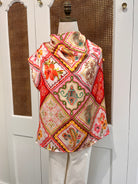 A folded textile item with a colorful patchwork-style floral and geometric print in red, pink, cream, green, and yellow, draped over a white chair in front of a white cabinet with woven inserts.