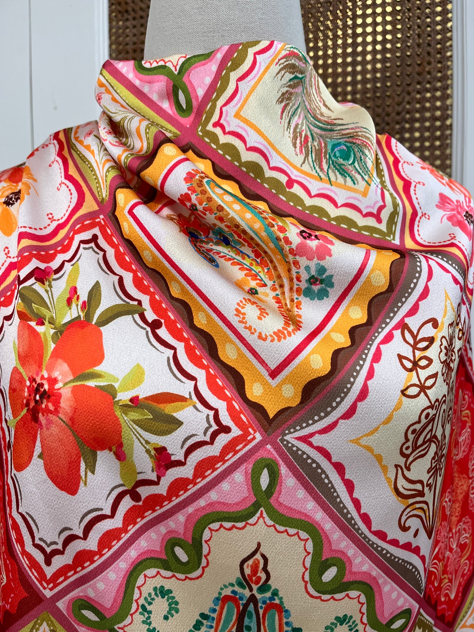 A close-up of a folded patterned fabric, likely a throw blanket or scarf, featuring a colorful patchwork design with floral, paisley, and peacock motifs in pink, orange, yellow, green, and brown on a white background.