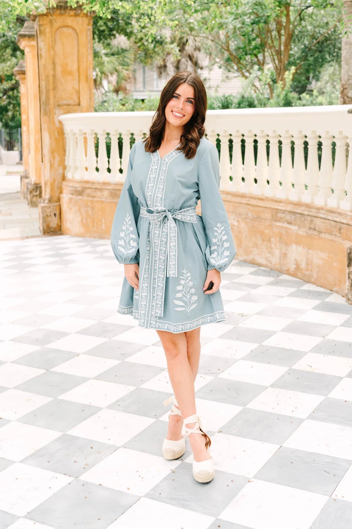 A woman wearing a light blue long-sleeve embroidered dress with a tie waist, standing on a black and white checkered outdoor patio.