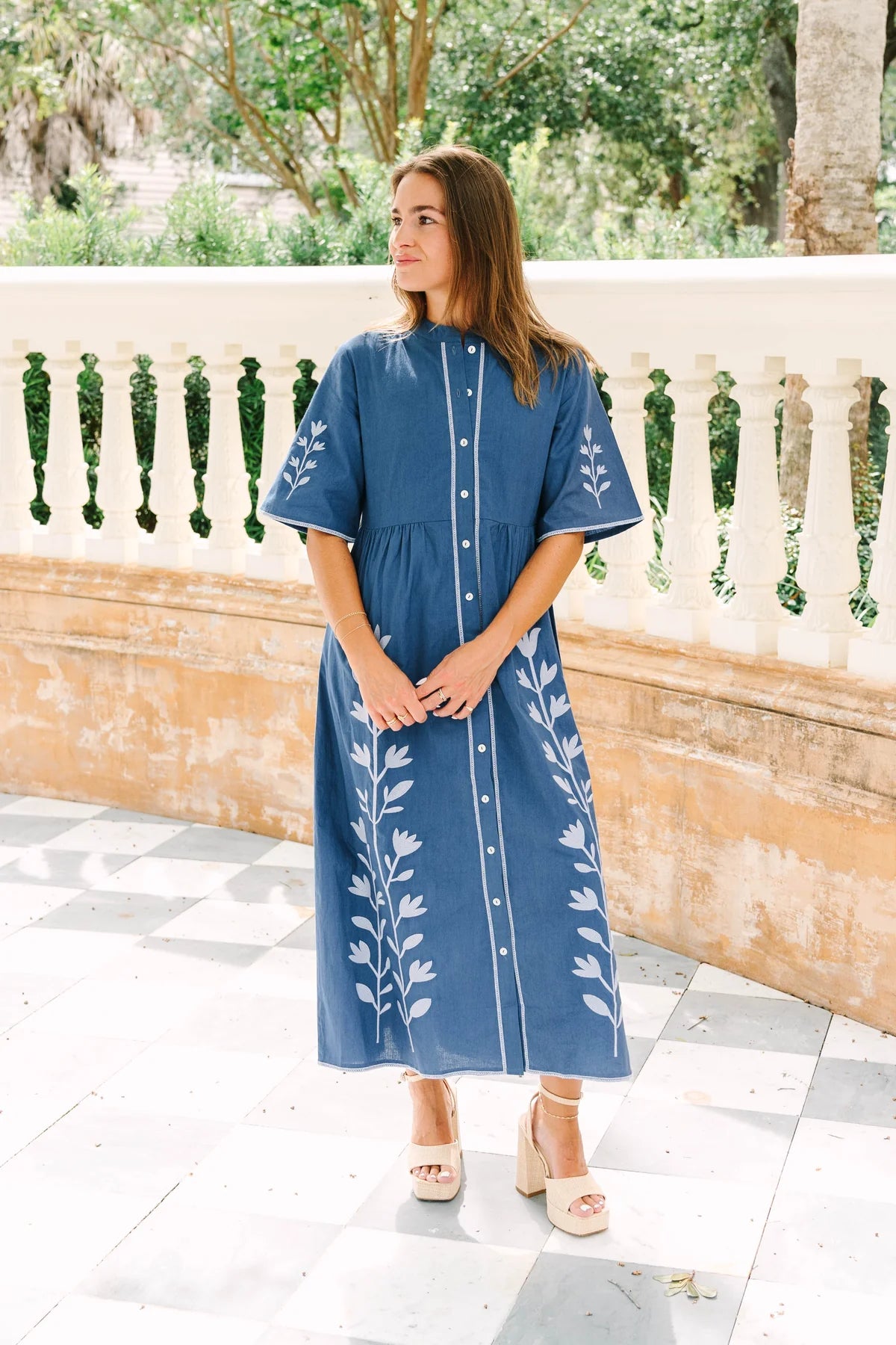 A woman wearing a blue midi dress with white floral embroidery and white buttons, standing on a tiled patio in front of a stone balustrade.