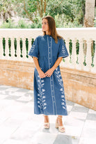 A woman wearing a blue midi dress with white floral embroidery and white buttons, standing on a tiled patio in front of a stone balustrade.